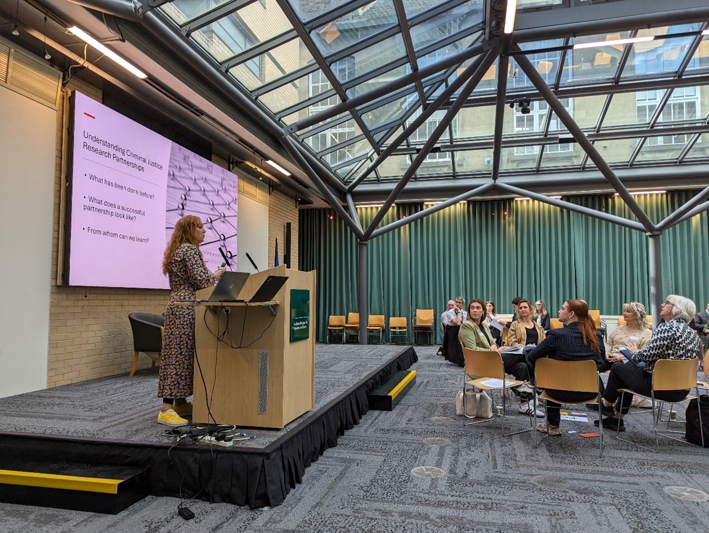 Patrice McCormack stands at a lectern in the department of justice meeting room, talking to audience members grouped around tables