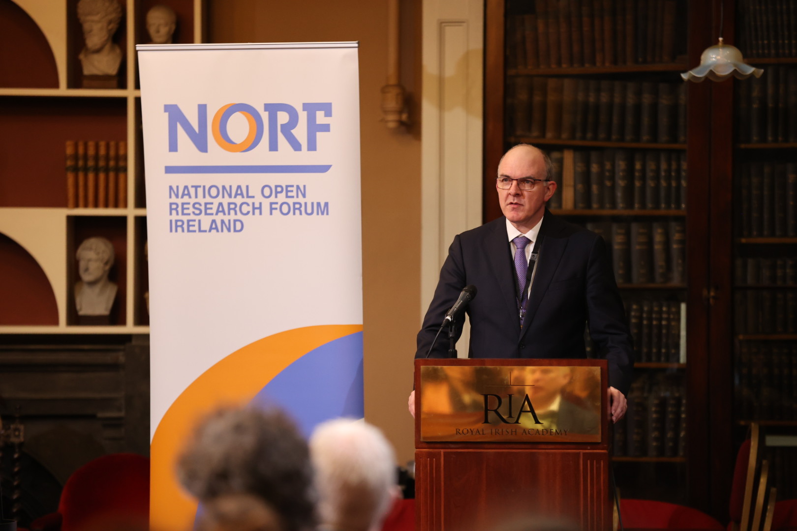 Opening remarks NORFest2023 A man in a suit and tie stands at a podium giving a speech at the Royal Irish Academy. A banner next to him reads "NORF - National Open Research Forum Ireland." The setting includes bookshelves and classical busts in the background, suggesting a formal academic environment.