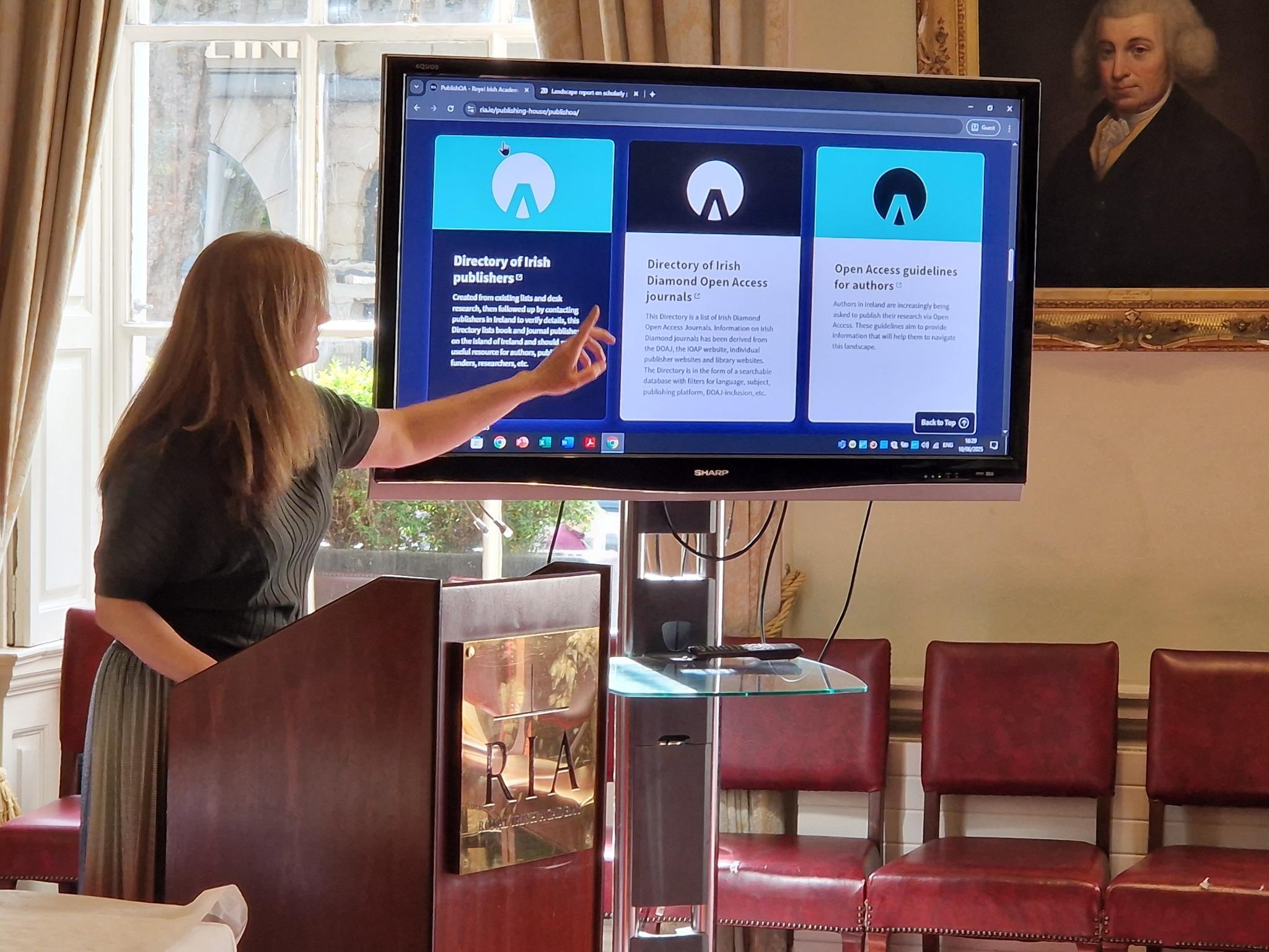Ruth Hegarty stands at a podium in a formal room, pointing at a large screen displaying a website with three panels. The website sections are titled "Directory of Irish publishers," "Directory of Irish Diamond Open Access journals," and "Open Access guidelines for authors." The room has elegant decor with red chairs, large windows, and a framed portrait hanging on the wall.