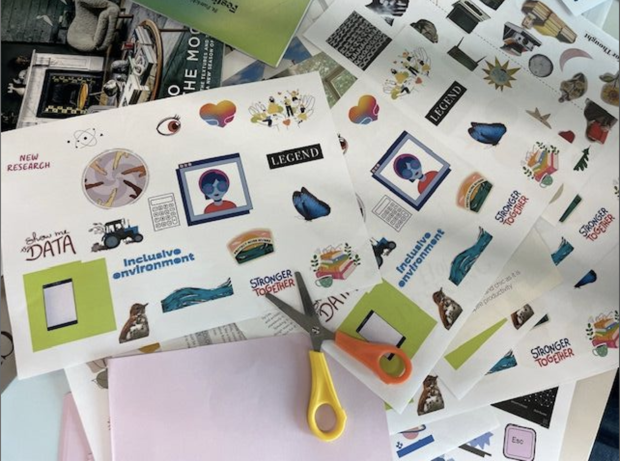 A cluttered workspace filled with sheets of printed stickers featuring various colorful illustrations and phrases like "Stronger Together," "Inclusive Environment," and "Show me my DATA." A pair of yellow and orange-handled scissors rests on the sheets, which include images of butterflies, abstract faces, sandwiches, and scientific graphics. Some magazines and green papers are partially visible underneath the sheets.