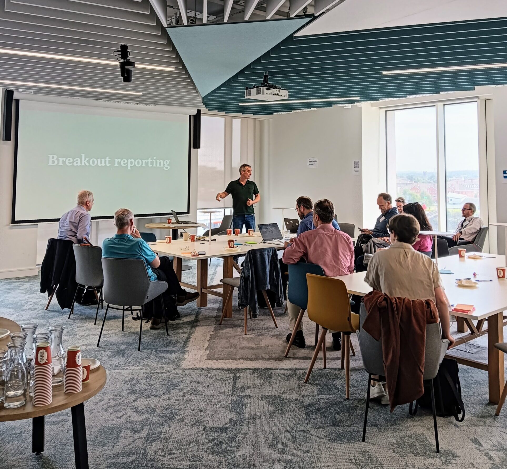 A group of people in a modern conference room listen to a speaker presenting in front of a screen that reads "Breakout reporting.