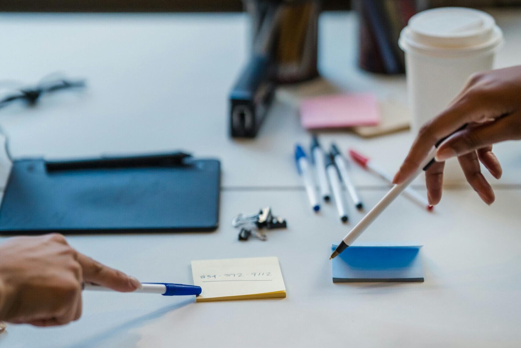 Two people point at sticky notes with pens on a desk cluttered with office supplies, a coffee cup, and writing tools near a window.