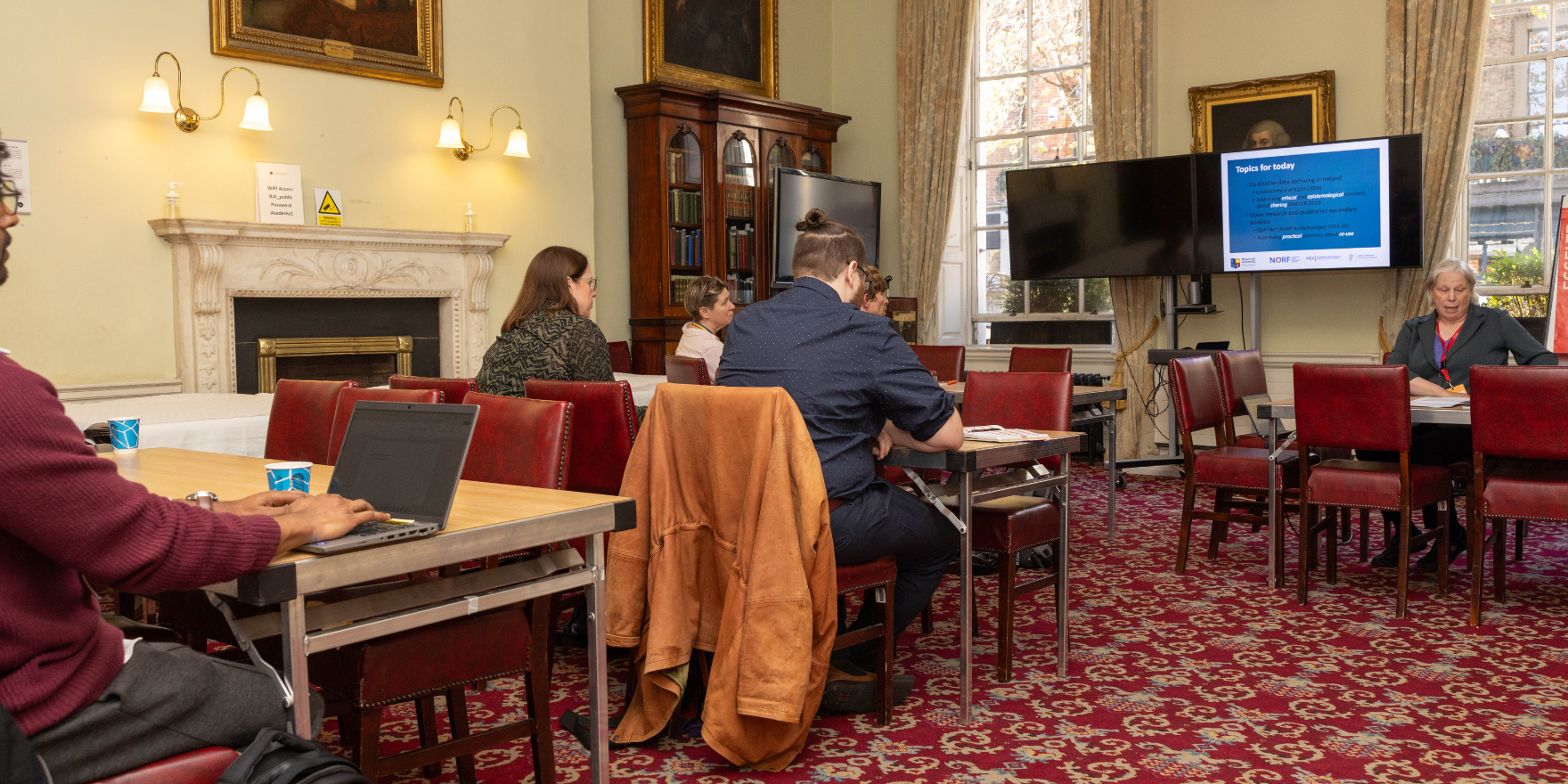 Individuals sitting at tables in the ornate Members' Room of the historic Royal Irish Academy building. They are at a workshop and have laptops, notes and a large screen at the top of the room.