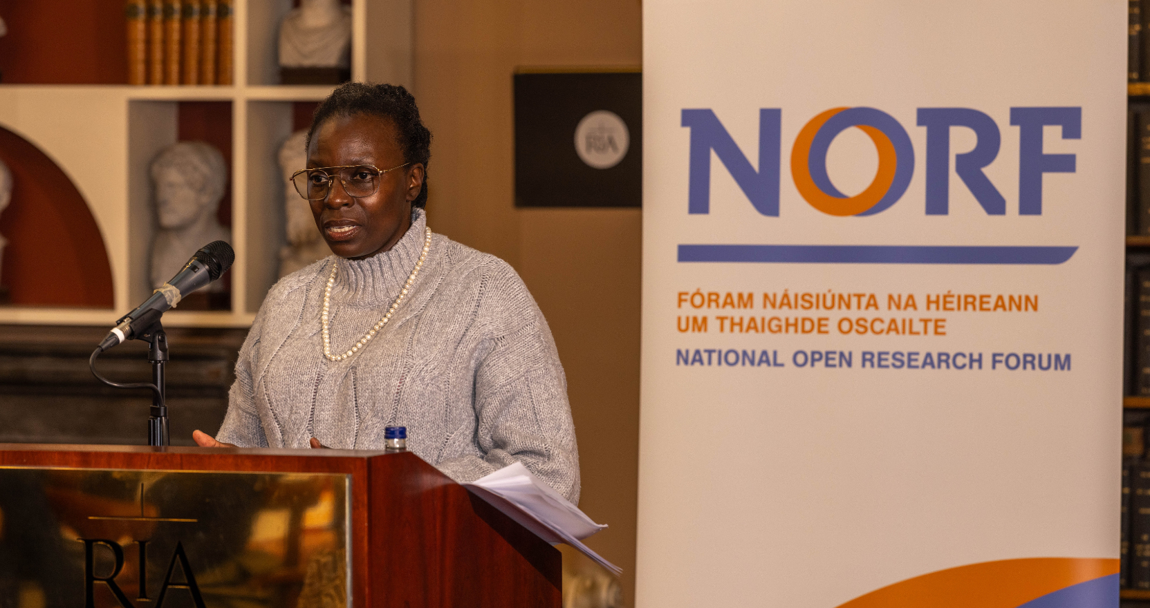 Ms Joy Owango standing at the podium in the historic Meeting Room of the Royal Irish Academy, delivering the keynote address at NORFest 2025. There is a banner in the background with the NORF bilingual logo on it. keynote speaker 2025.