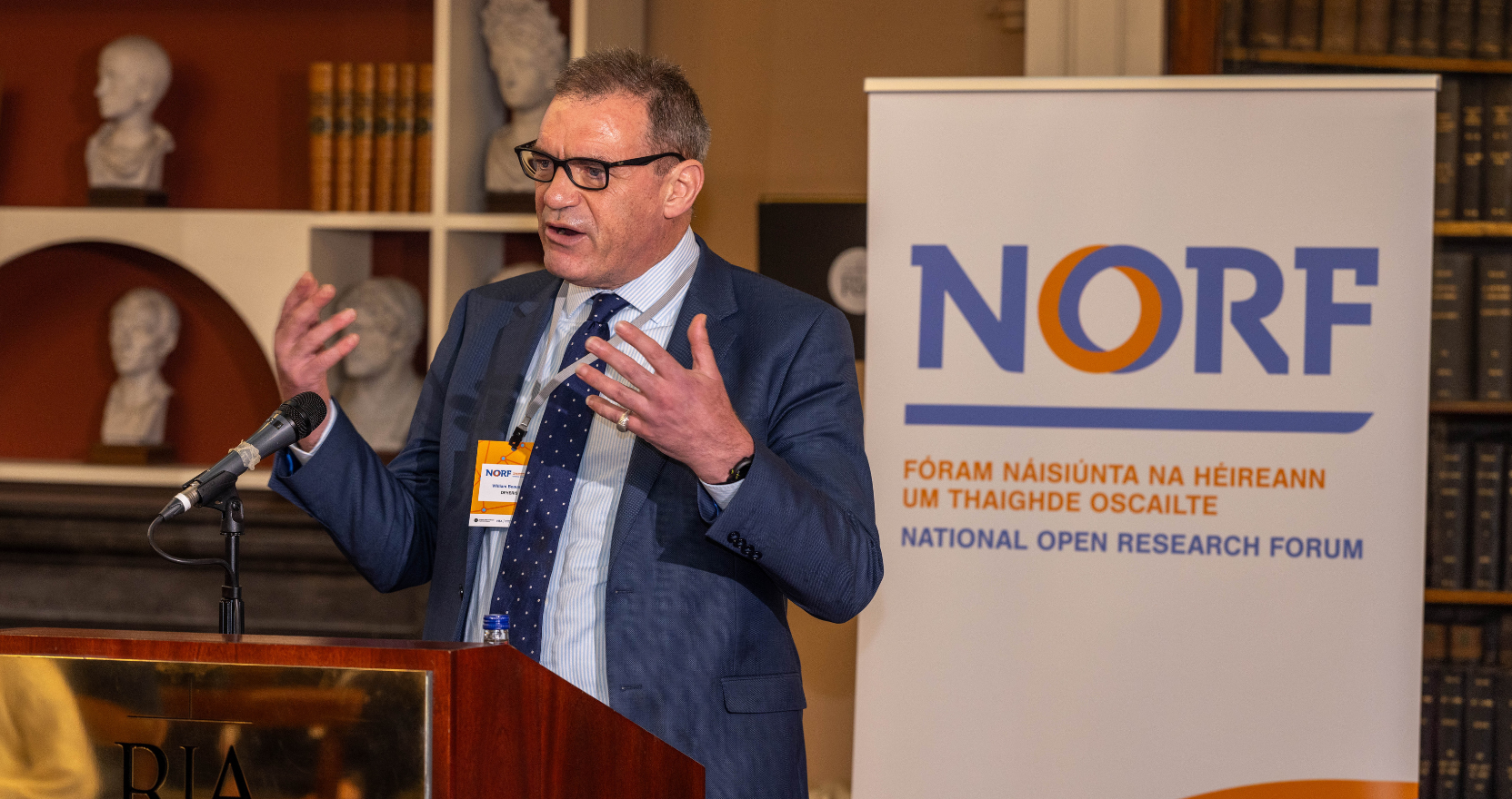 A man in a suit and tie stands at a podium giving a speech at the Royal Irish Academy. A banner next to him reads "NORF - National Open Research Forum Ireland." The setting includes bookshelves and classical busts in the background, suggesting a formal academic environment.