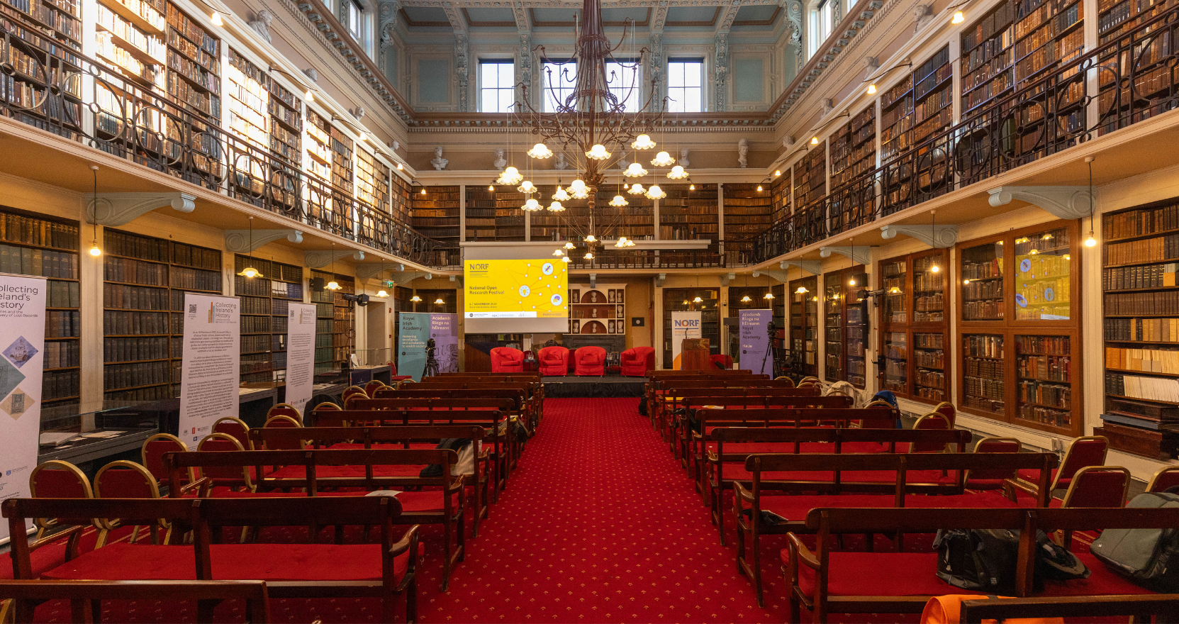 The Meeting Room of The Royal Irish Academy showing its historic architecture and library collection. Rows of empty benches face a stage and podium.