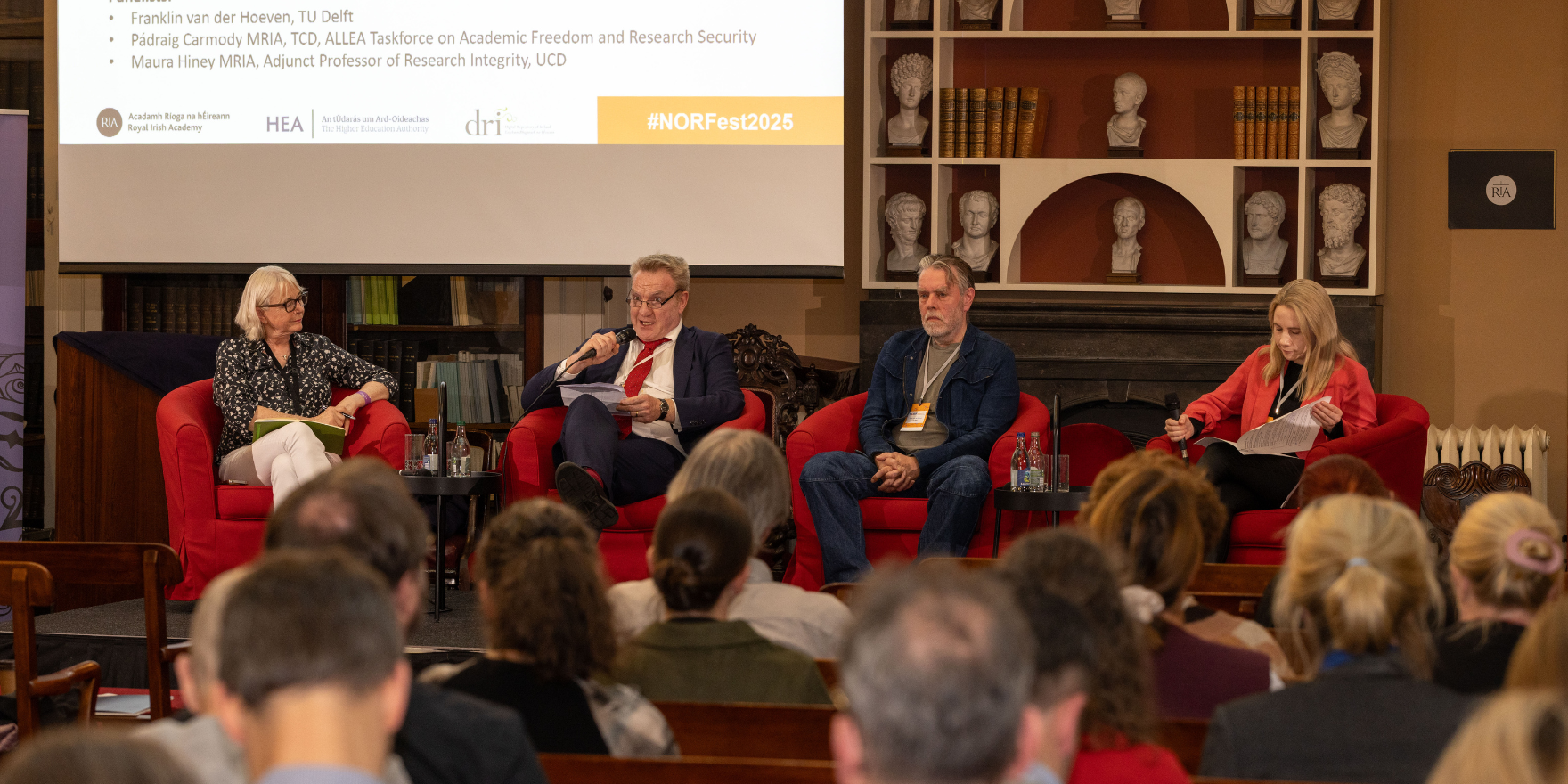 Maura Hiney MRIA, Pádraig Carmody MRIA, Franklin van der Hoeven and Senator Laura Harmon sitting on red chairs on a stage in the Meeting Room of the Royal Irish Academy, participating in a panel discussion during NORFest 2025 Day One. They are holding hand held microphones and a large projector screen is above their heads to the left.