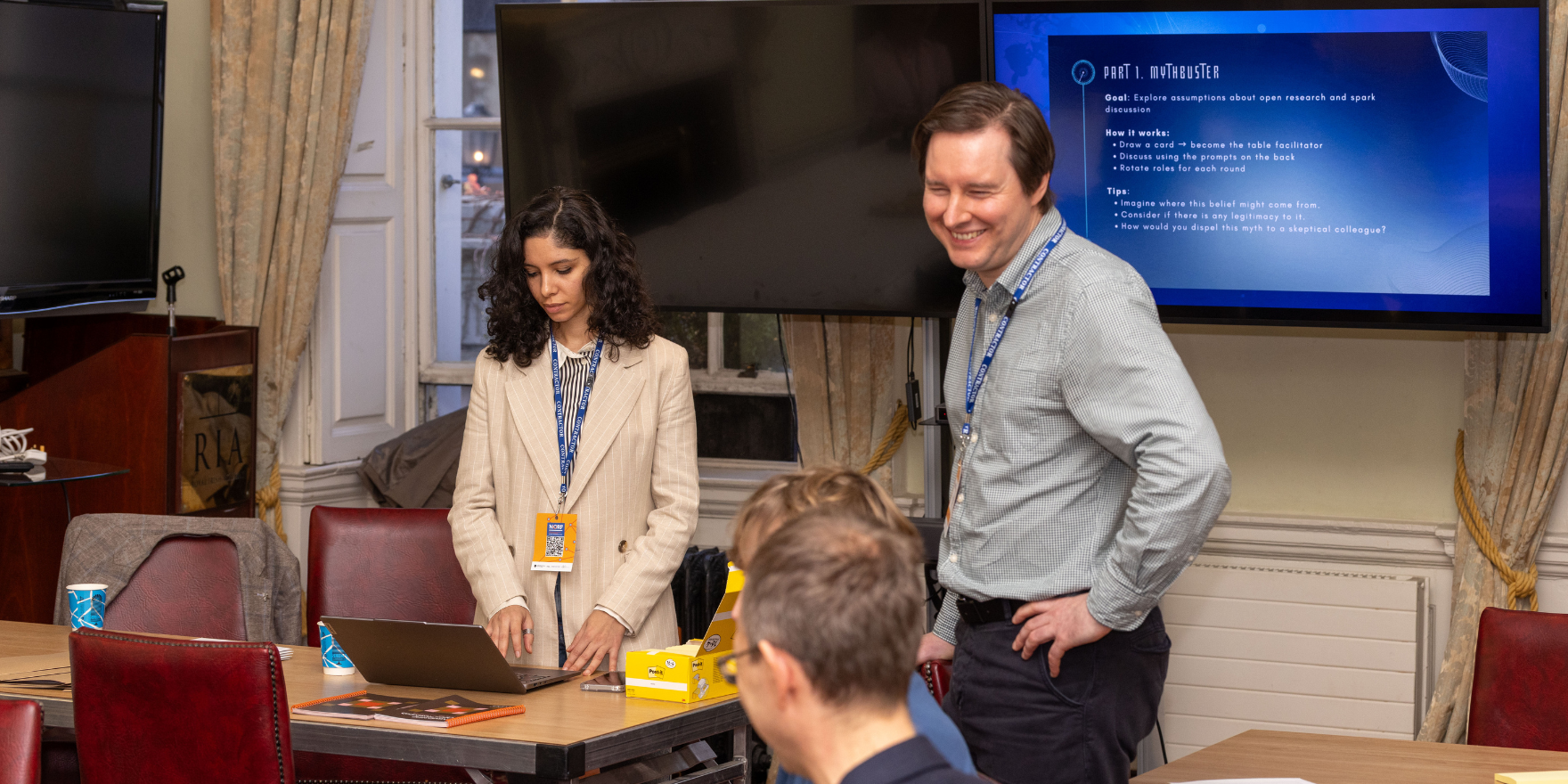 Two individuals sitting at a table in the ornate Members' Room of the historic Royal Irish Academy building. They are at a workshop hosted by Dounia Lakhzoum who is standing up looking at her laptop with a large screen behind her. Dermott Lynott is standing next to her, chatting to the participants and smiling.