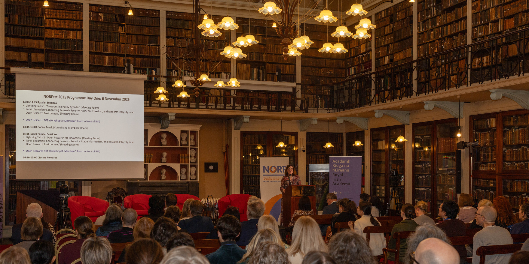 Dr Michelle Doran, National Open Research Coordinator, speaking from the podium in the Meeting Room of the Royal Irish Academy showing its historic architecture and library collection. Rows of benches are filled with event attendees.