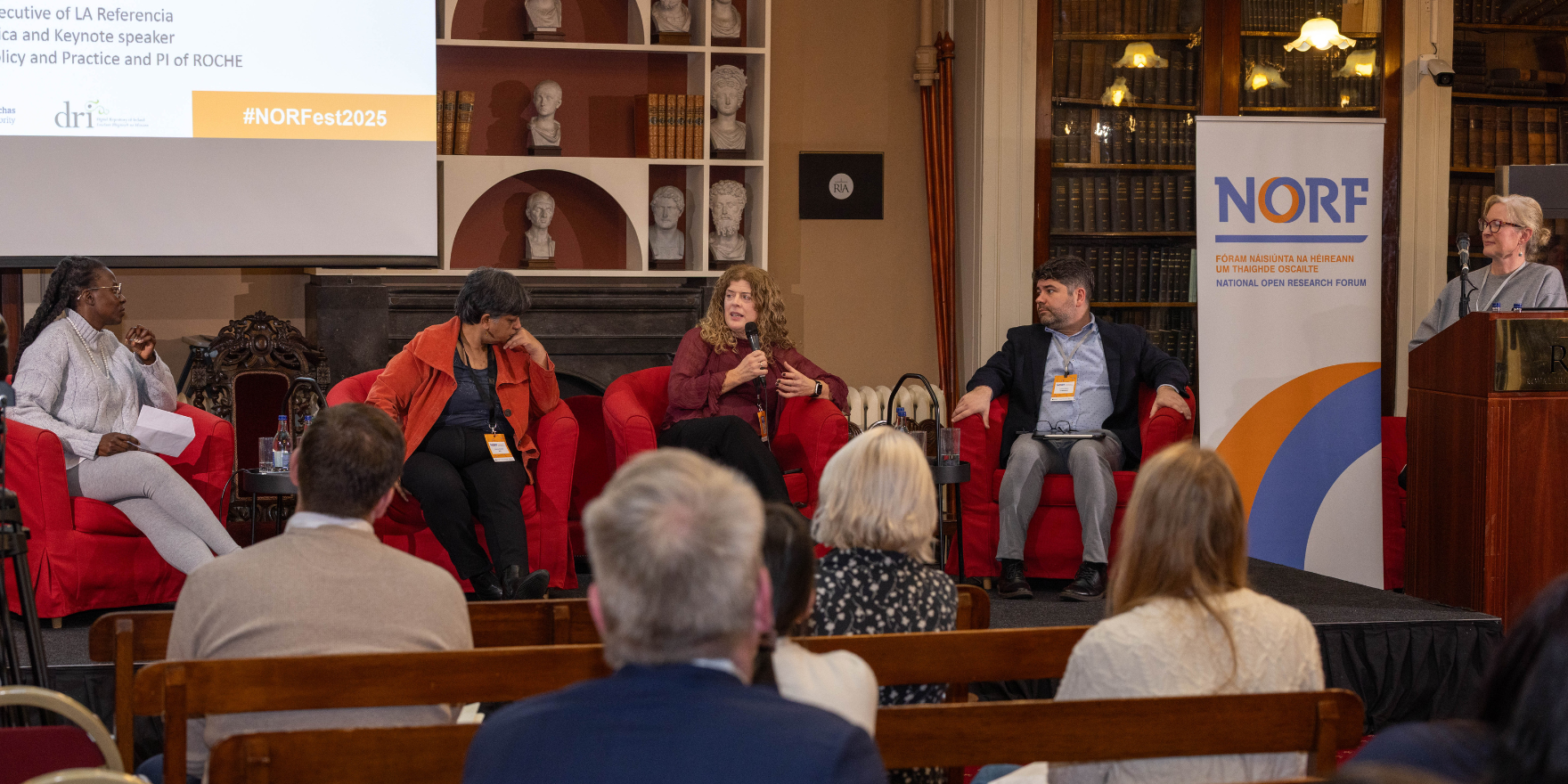Joy Owango, Kalpana Shankar, Aedin Culhane and Lautaro Matas sitting on red chairs on the stage in the historic Meeting Room of the Royal Irish Academy, participating in a panel discussion during NORFest 2025 Day One. Clare Dillon who is chairing the panel stands at the podium to the right of the stage.