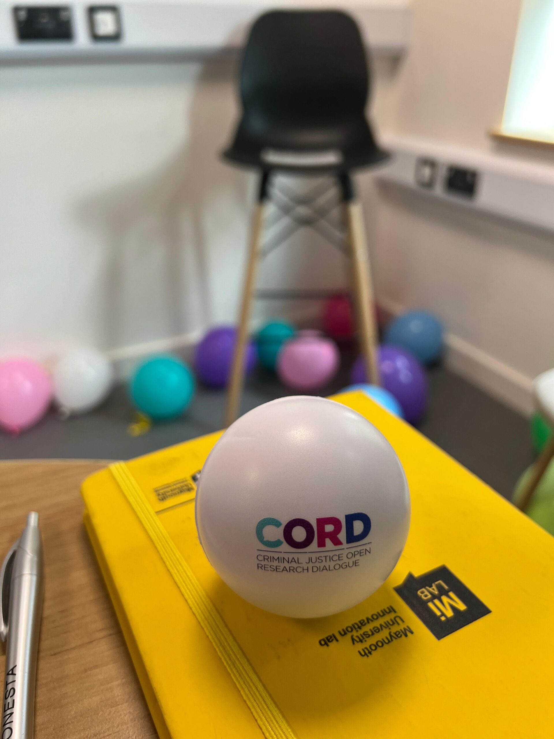 White stress ball with CORD logo set upon a yellow book. The background features multiple colourful balloons.